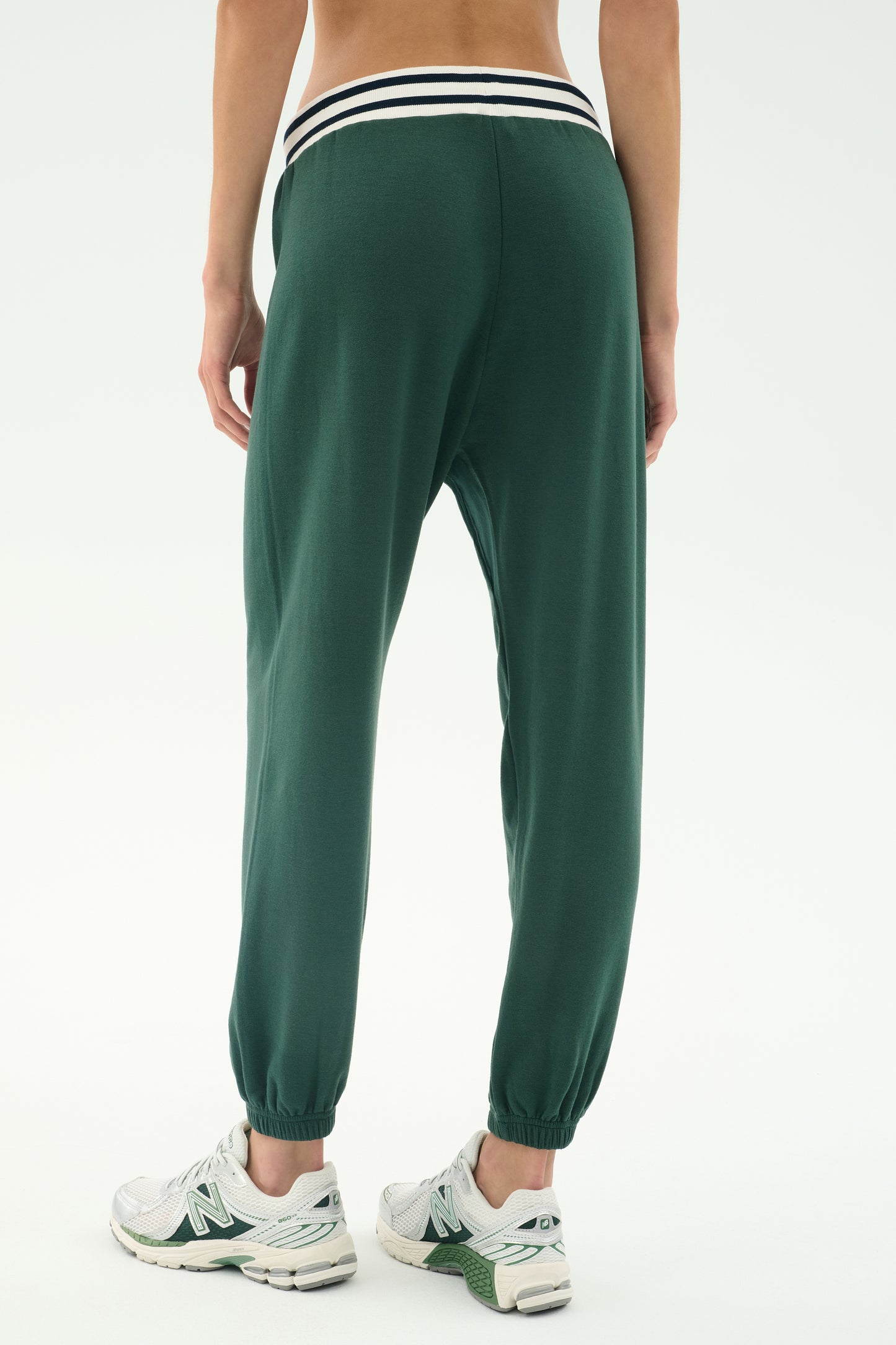 A person shown from the back wears SPLITS59’s Andie Oversized Fleece Sweatpant W/ Stripe in Military with a striped waistband and white athletic shoes, standing against a plain background.