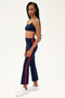 A woman stands sideways in a SPLITS59 Raquel High Waist Crop in Indigo/Pirate Red, featuring 4-way stretch supplex fabric and red side stripes, paired with matching flared pants and white sneakers against a plain background.
