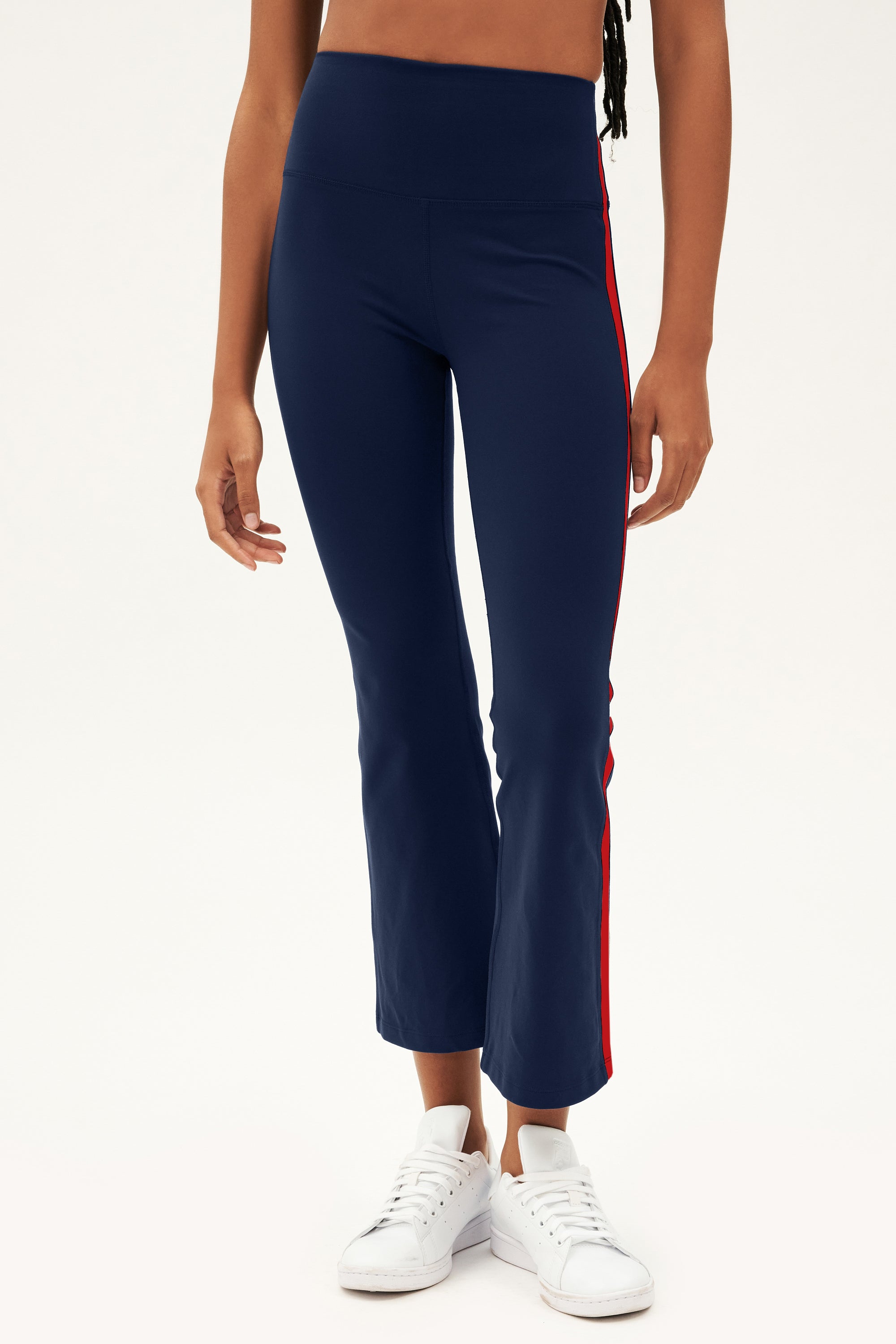 A person wears SPLITS59 Raquel High Waist Crop pants in Indigo/Pirate Red, made from 4-way stretch supplex, with white sneakers, shown from shoulders down against a plain background.