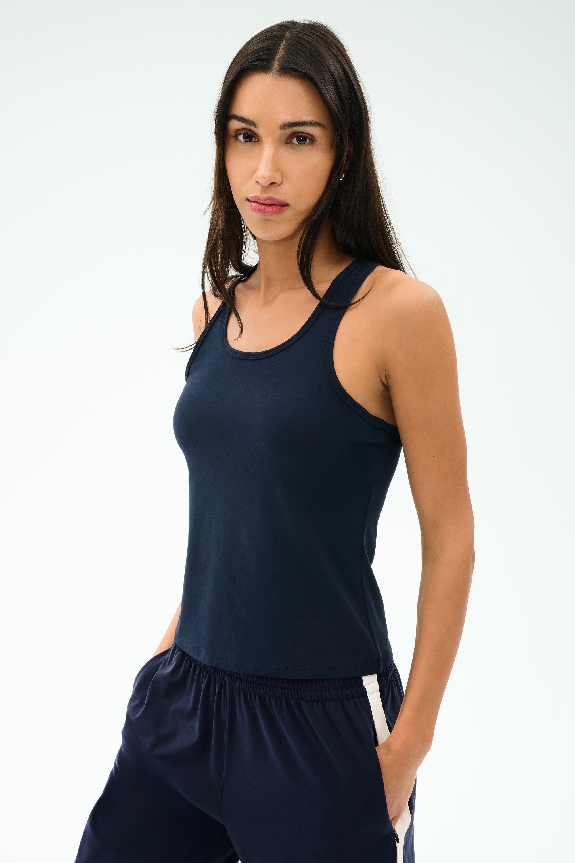 A woman wears the SPLITS59 Mel Airweight Lite Tank in Indigo, paired with matching athletic pants, standing hands-in-pockets against a light background.