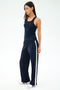 A woman models the SPLITS59 Mel Airweight Lite Tank in indigo and matching navy track pants with white side stripes, standing sideways against a plain white background.