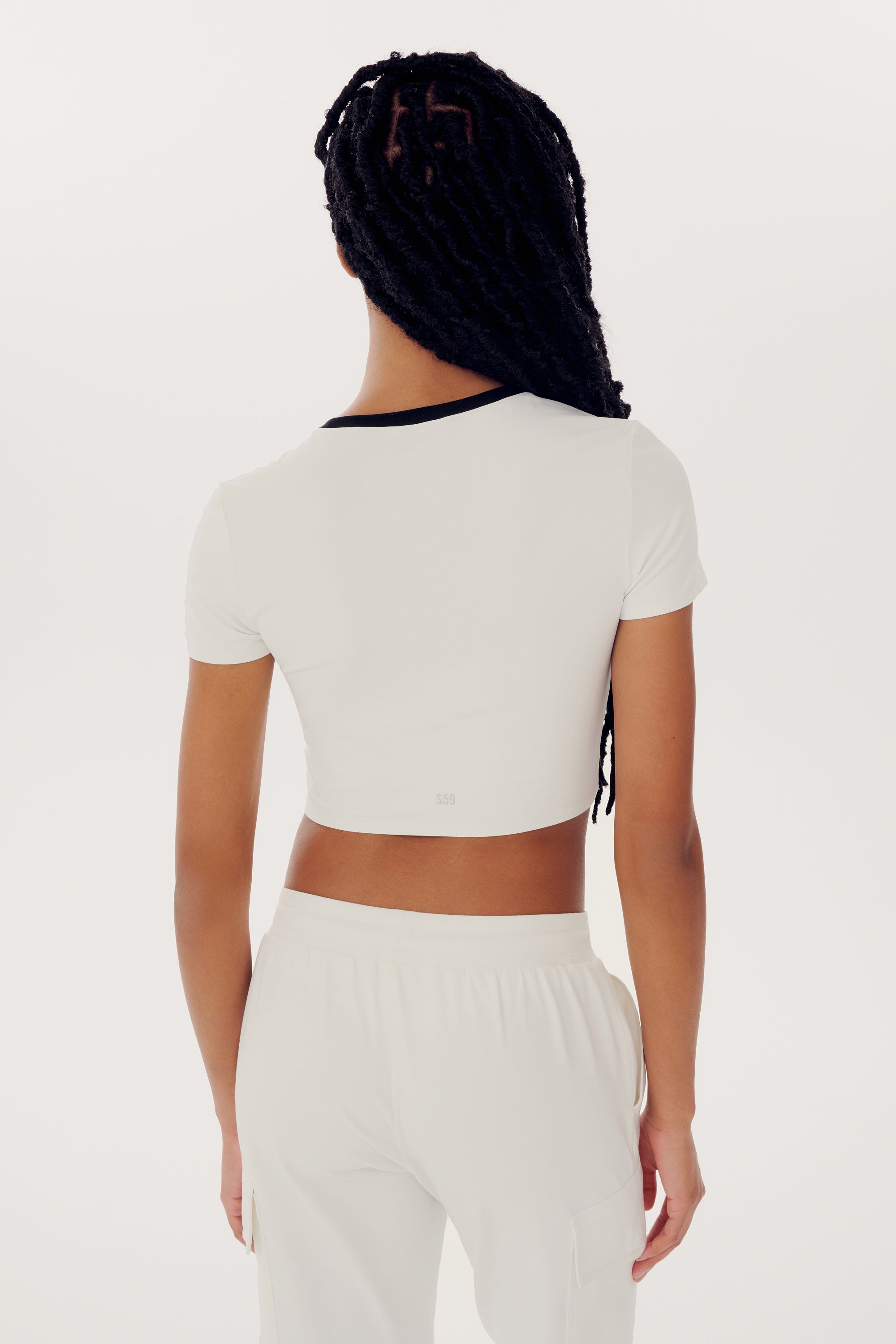 A person wearing a white short-sleeved Airweight S/S Crop - White/Black by SPLITS59 and white nylon pants is standing with their back facing the camera. They have long braided hair.