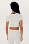 A person wearing a white short-sleeved Airweight S/S Crop - White/Black by SPLITS59 and white nylon pants is standing with their back facing the camera. They have long braided hair.