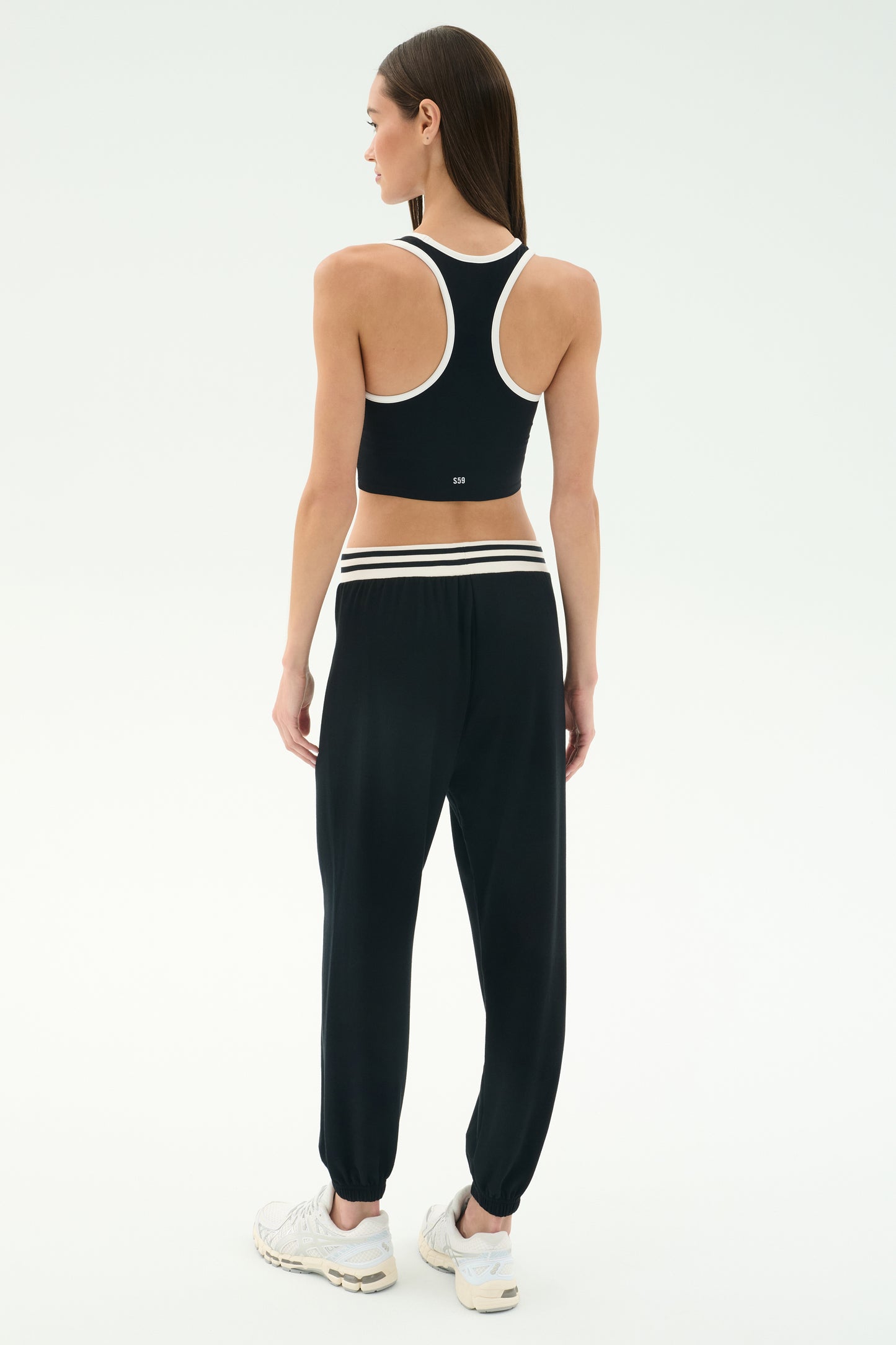 A woman faces away, wearing SPLITS59 Andie Oversized Fleece Sweatpant W/ Stripe in black and a black racerback sports top, paired with white sneakers against a light background.