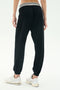Person wears the SPLITS59 Andie Oversized Fleece Sweatpant W/ Stripe in black, featuring a white stripe waistband, paired with white sneakers and shown from the back against a plain white background.