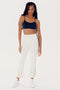 A woman is standing confidently in a black sports bra, high waist SPLITS59 Raquel High Waist Crop - White/Indigo pants with side stripes made of 4-way stretch supplex fabric, and matching white sneakers. She has long braided hair and is posing against a plain white background.