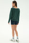 A woman stands with her back to the camera, wearing the SPLITS59 Sonja Fleece Sweatshirt W/ Stripe in Military, paired with navy shorts, white socks, and sneakers against a plain background.