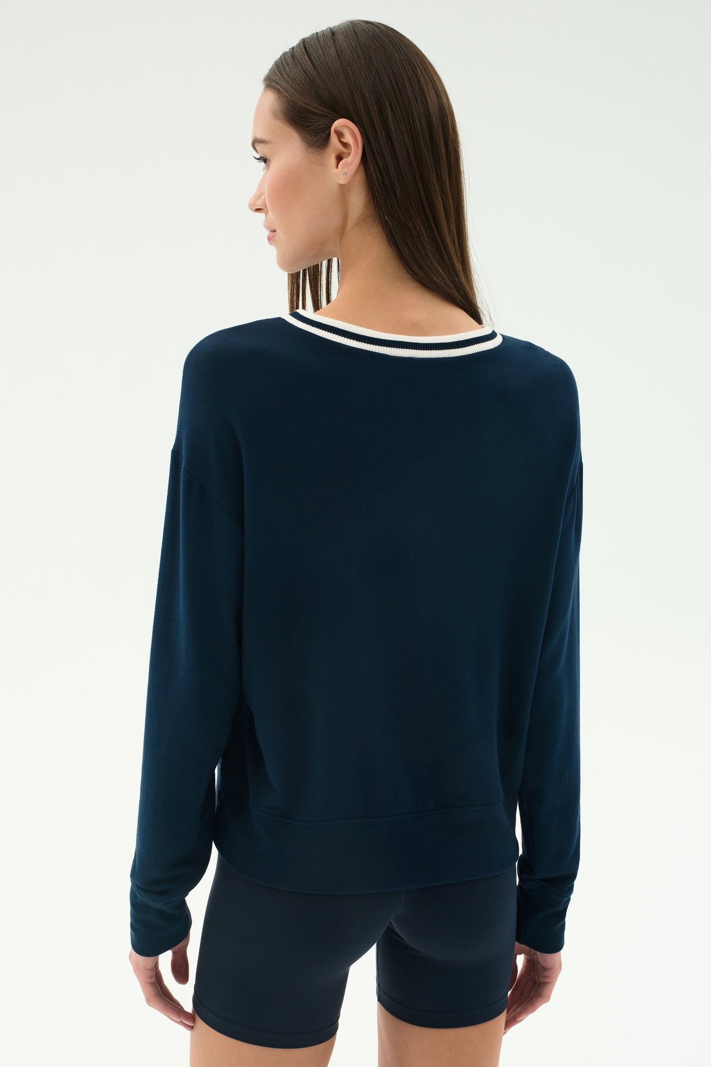 A woman with straight brown hair faces away, wearing the SPLITS59 Sonja Fleece Sweatshirt W/ Stripe in indigo and matching shorts, set against a plain light background.