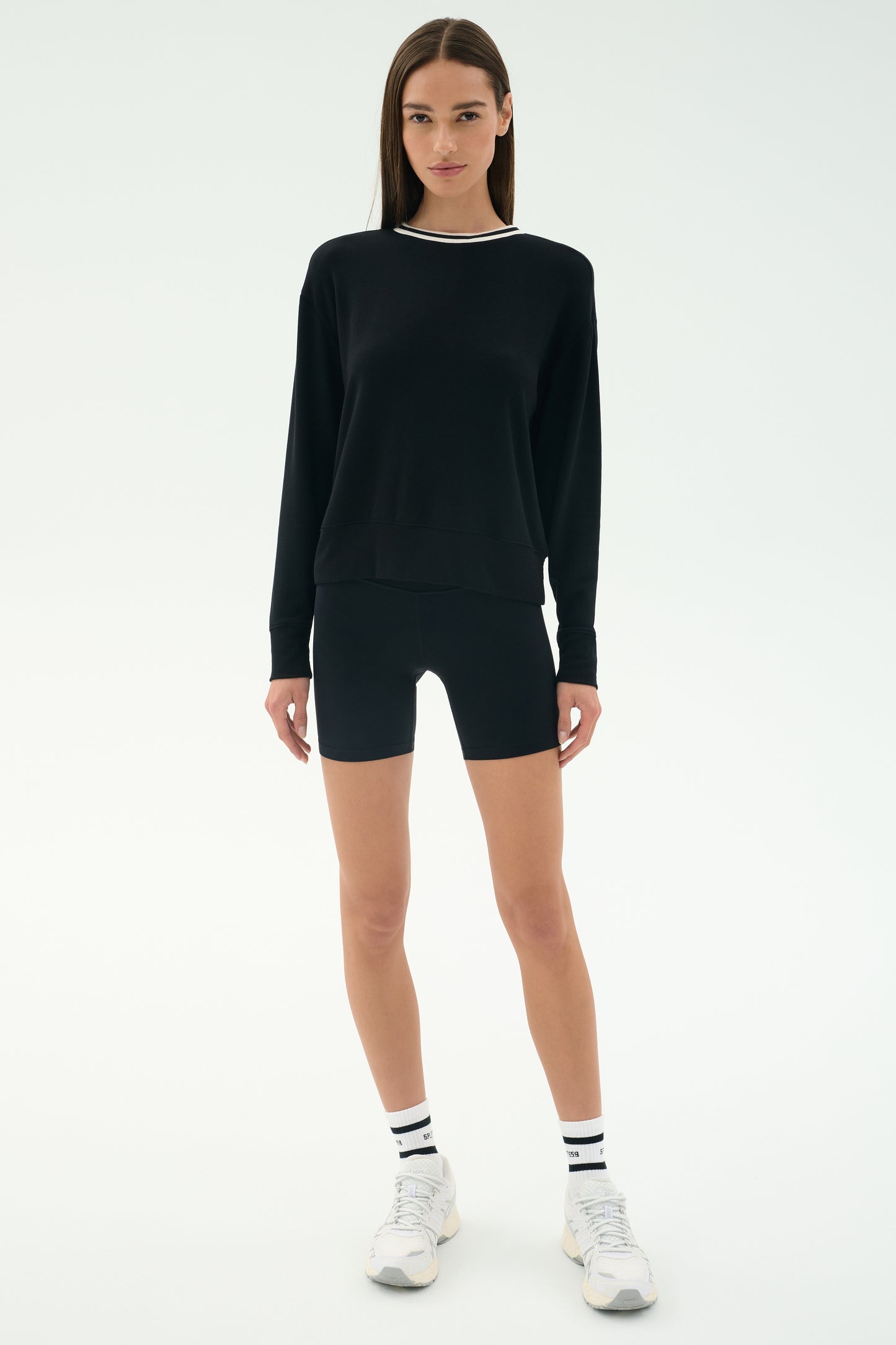 A woman faces forward on a plain light background wearing the SPLITS59 Sonja Fleece Sweatshirt W/ Stripe, black fitted shorts, white sneakers, and white socks with black stripes.