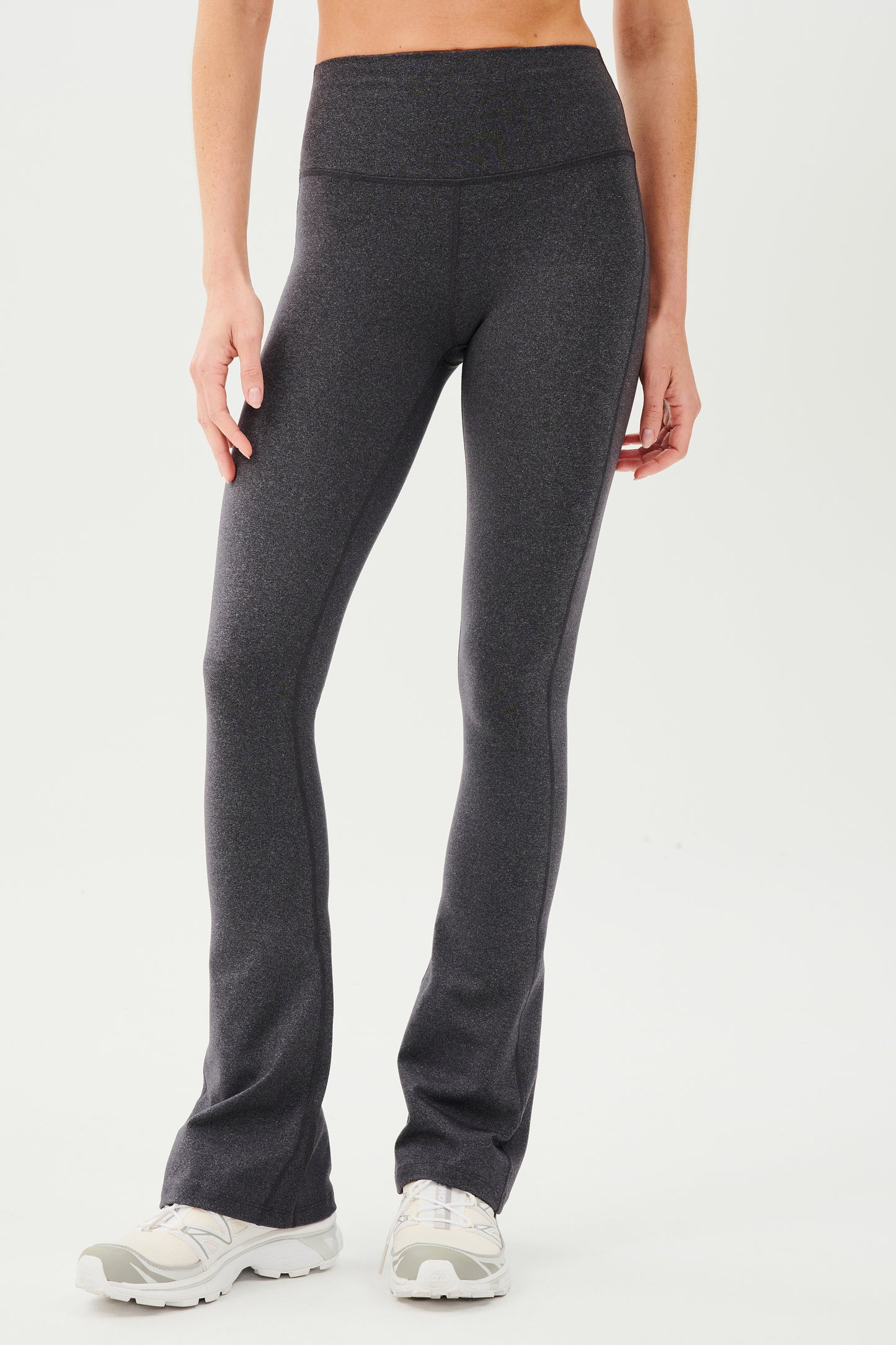 Front view of woman wearing dark gray high waist below ankle length legging with wide flared bottoms. Paired with white shoes.