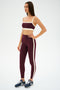 A woman stands in a studio wearing the SPLITS59 Clare High Waist Rigor 7/8 leggings in Dark Cherry/Creme with a matching sports bra and navy blue athletic shoes.