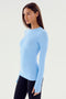 A person with long dark hair is wearing a form-fitting Skylight Louise Rib Long Sleeve from SPLITS59 and black leggings, standing against a plain white background.
