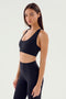 A person with long hair is wearing a SPLITS59 Sara Airweight Bra in black and black high-waisted leggings, standing against a plain white background, ready for a Pilates session.
