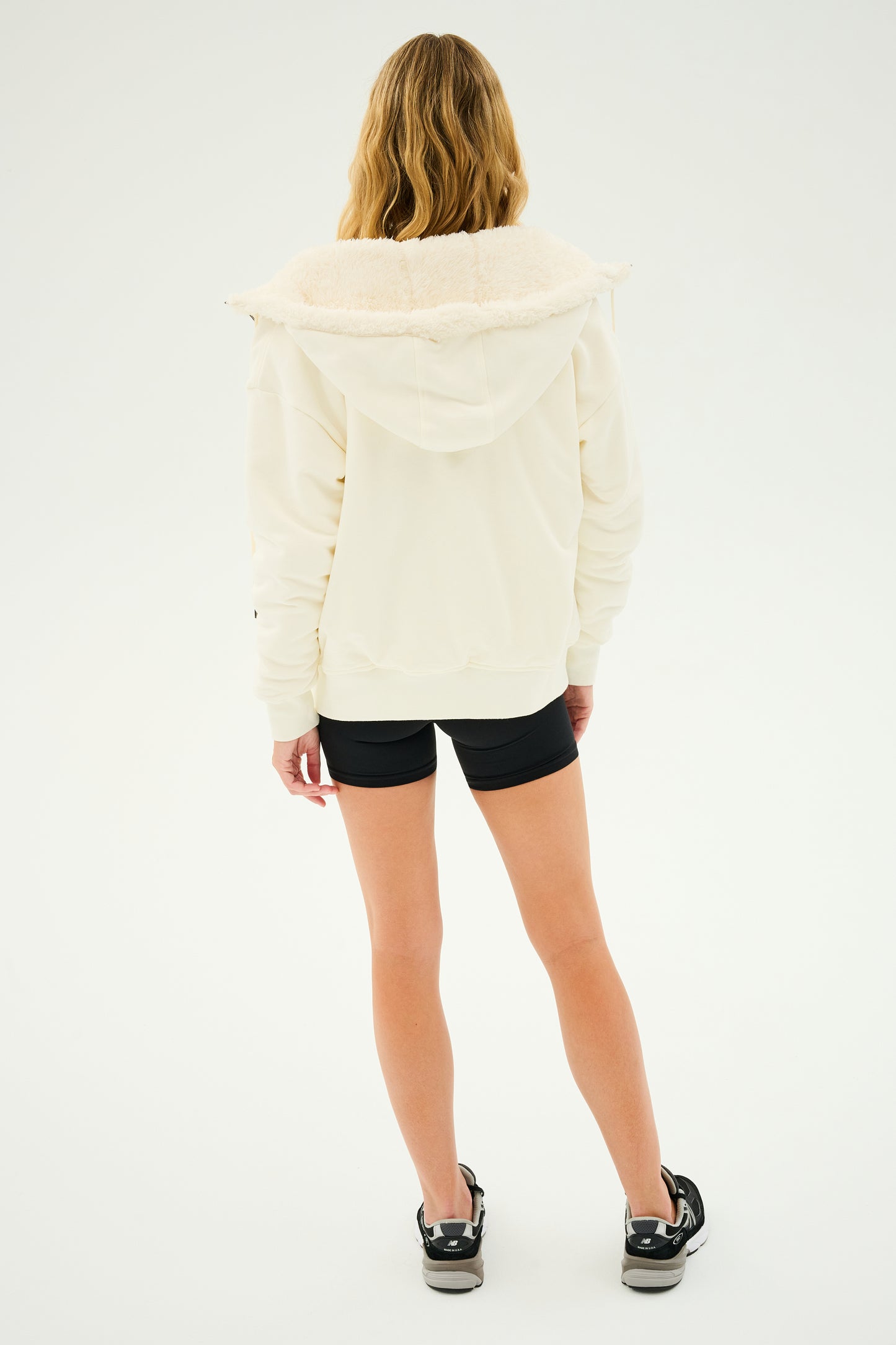 A person with long hair, dressed in a cozy Lynx French Terry Sherpa Hoodie by SPLITS59 in Creme/Creme, black shorts, and black sneakers, stands with their back to the camera against a plain background.
