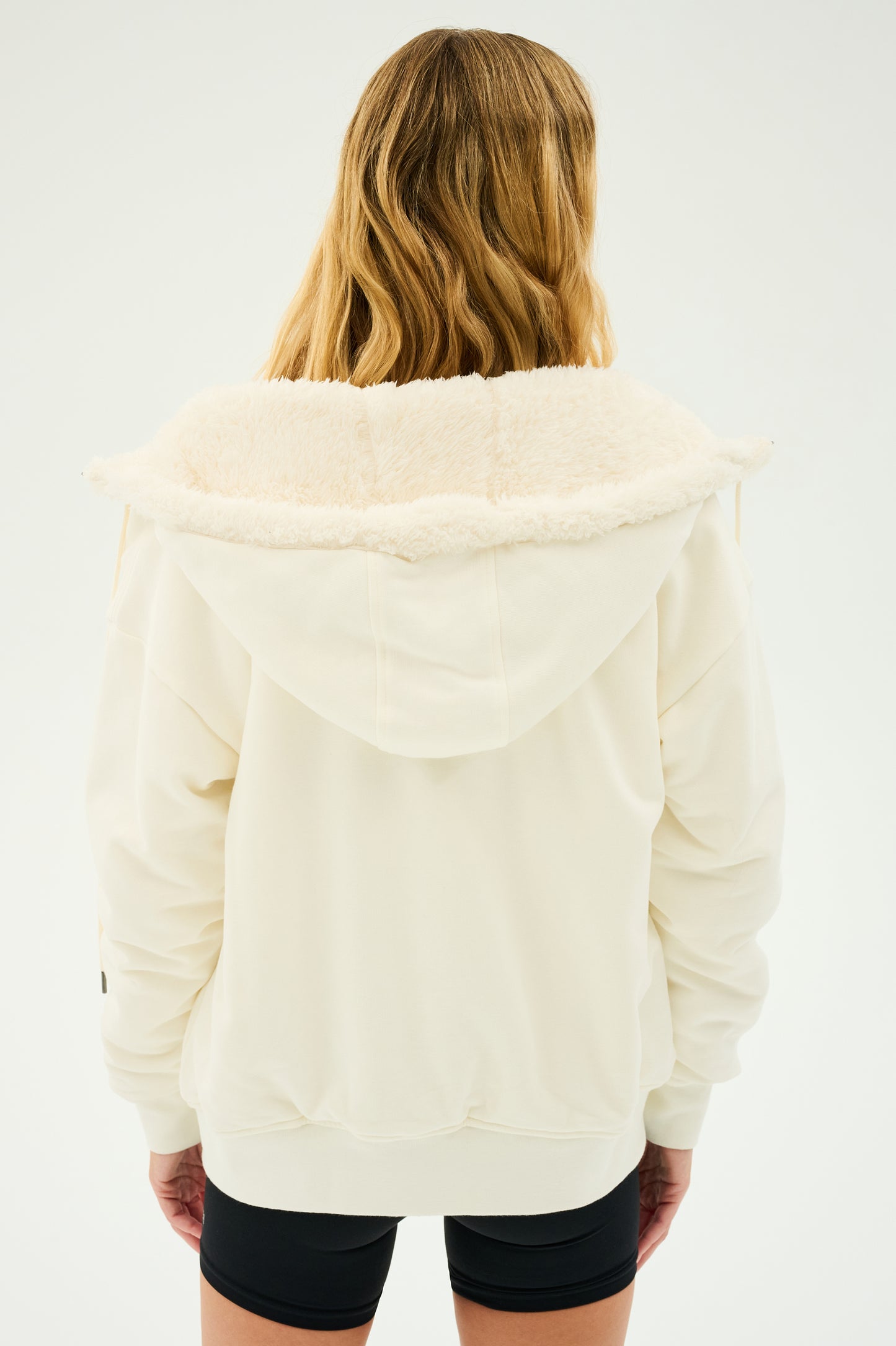 A person with long hair, seen from behind, is wearing the SPLITS59 Lynx French Terry Sherpa Hoodie in Creme/Creme, perfect for après-ski, complemented by black shorts.
