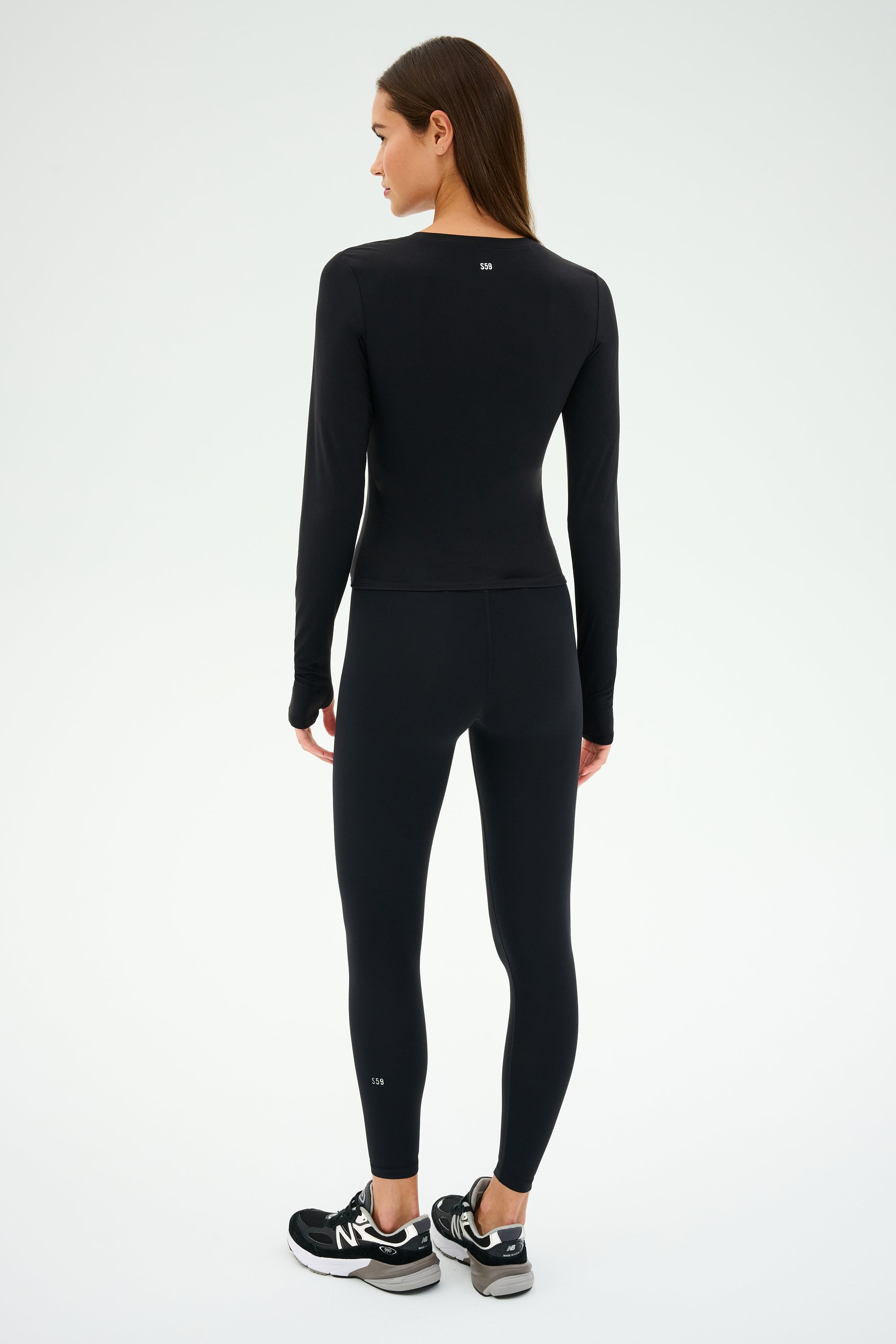 Dressed in the Airweight Lite LS Waist Length - Black by SPLITS59, an individual in sleek athletic wear and quick-dry sneakers stands on a white background, prepared for intense gym workouts.