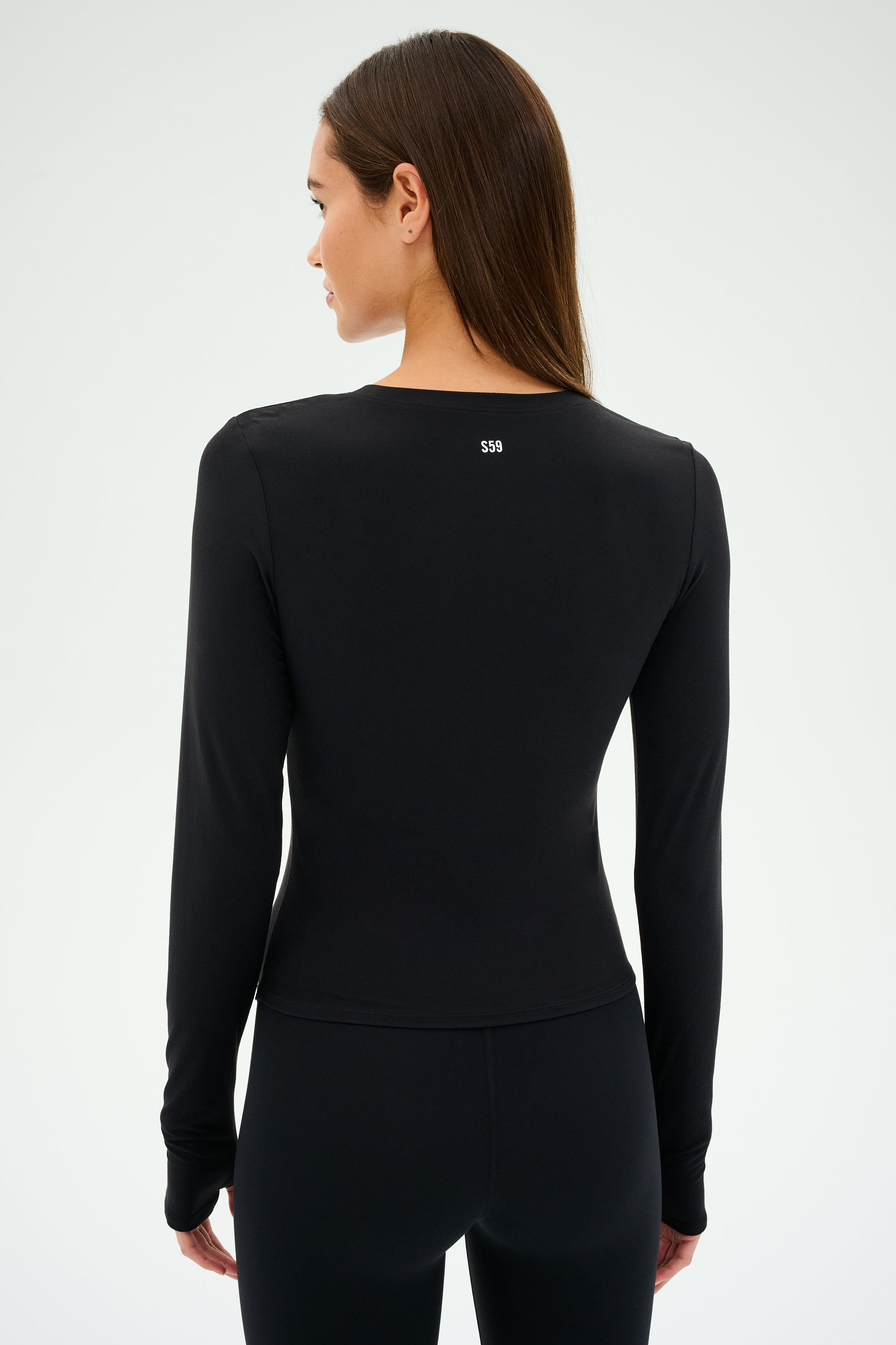 A person wears an SPLITS59 Airweight Lite LS Waist Length top and pants in black, facing away with a small white logo on the upper back. Crafted from airweight fabric, it's ideal for gym workouts against a plain, light background.