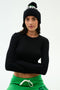 Woman wearing a black long-sleeve top, green drawstring pants, and a black beanie with a pom-pom, standing with arms crossed against a plain background.