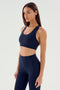 A person with long, straight hair is wearing a matching dark blue Sara Airweight Bra by SPLITS59 along with high-waisted leggings. This outfit is perfect for hot yoga or Pilates. They are standing against a plain, light-colored background.