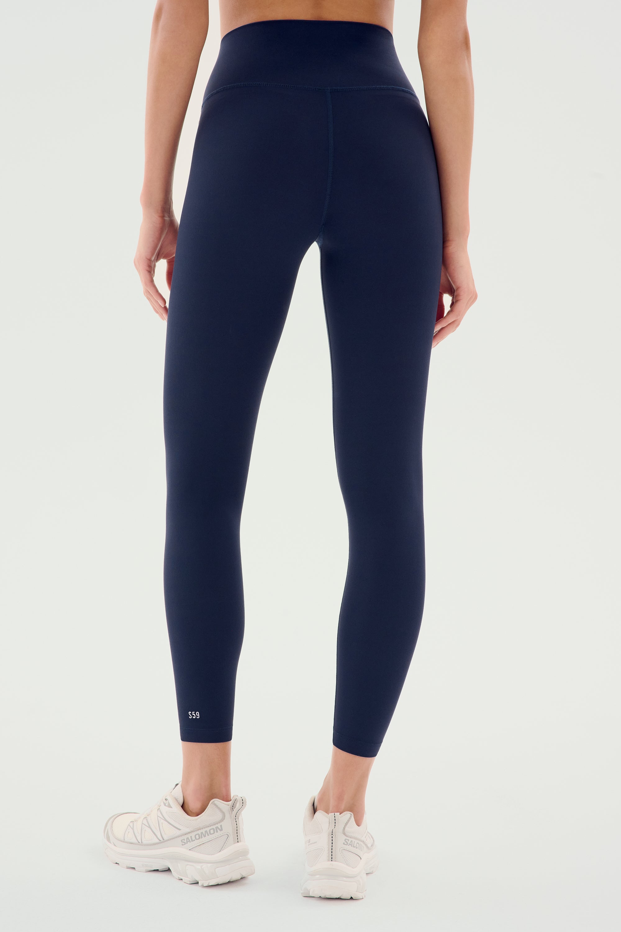 A person is wearing SPLITS59's Airweight High Waist Legging in Indigo paired with white sneakers, shown from the back against a plain background. Perfect for hot yoga sessions.