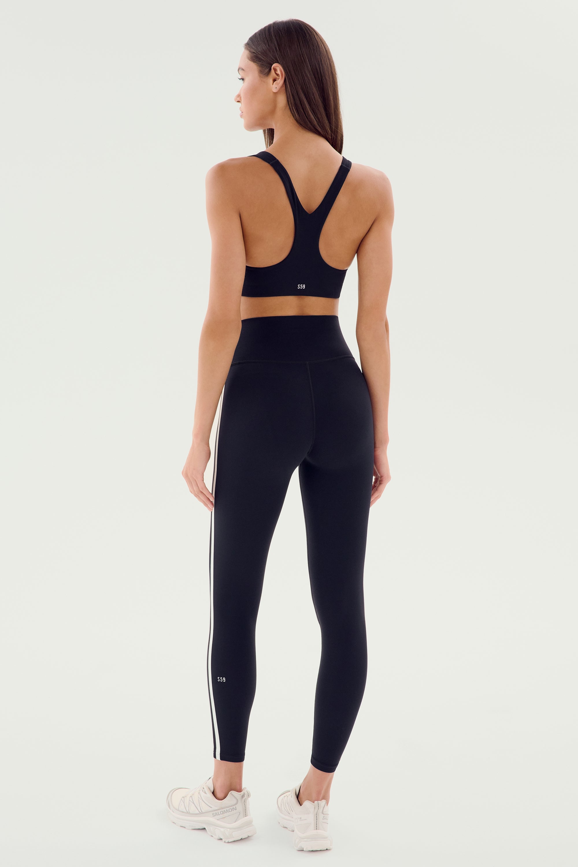 A woman faces away, wearing the SPLITS59 Ella High Waist Airweight 7/8 leggings in Black/White and a matching sports bra, paired with white sneakers against a plain light background.