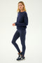 A person stands in profile against a plain light background, wearing the SPLITS59 Sonja Fleece Sweatshirt in Indigo, matching leggings, and black athletic shoes.