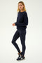 A person stands against a plain background wearing the SPLITS59 River High Waist Airweight Stirrup leggings in Indigo, a matching navy long-sleeve top, and black athletic shoes.