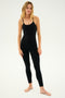 A woman stands barefoot, wearing the SPLITS59 Airweight Jumpsuit - Black, a body-hugging piece made from Airweight fabric with thin straps. She poses facing the camera against a plain white background.