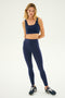 A woman is standing against a plain white background, modeling her Indigo Sprint Rigor Bra by SPLITS59, matching leggings, and black sneakers, ready for gym workouts and multi-sport performance.