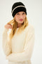 A person with long blonde hair, wearing the SPLITS59 Rib Beanie in Black/Creme and a cozy cream-colored sweater, looks directly at the camera.