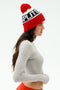 Woman in a SPLITS59 Winter Pom Pom Beanie—red, white, and navy with "SPLITS" text—gray long-sleeve shirt, and red pants, stands in profile against a plain background.