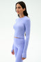 A woman stands against a plain light background wearing SPLITS59's Louise Rib Long Sleeve Crop in Purple Haze with matching high-waisted leggings.