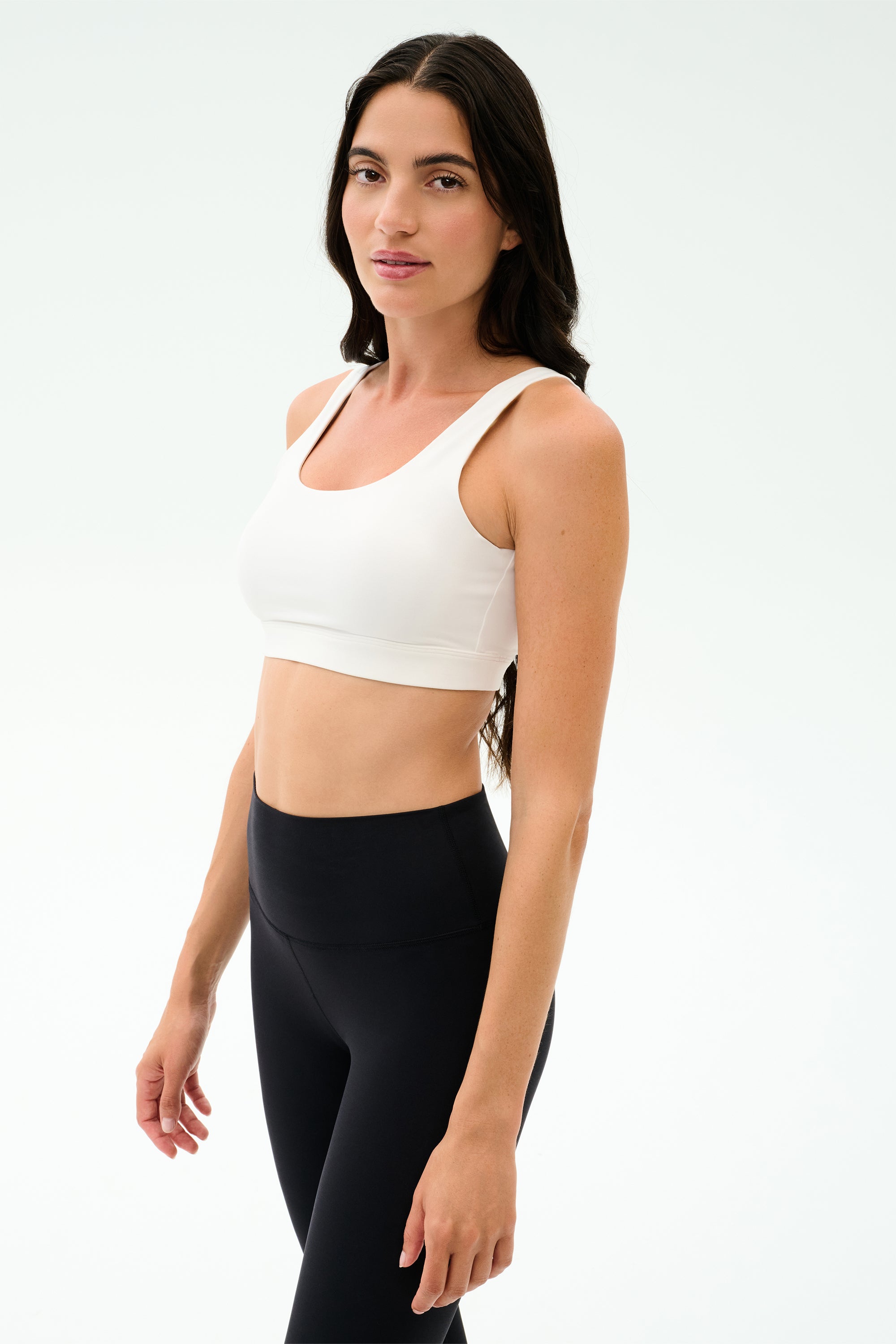 Woman with long dark hair wears the SPLITS59 Sprint Rigor Bra in white and black high-waisted leggings, standing in a well-lit studio with a neutral background—perfect for gym sessions and versatile multi-sport performance.