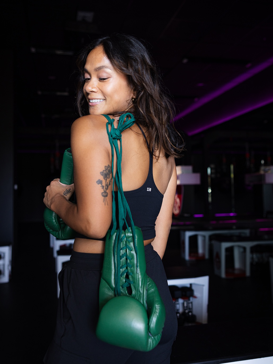A woman in athletic wear stands indoors, smiling over her shoulder, with green boxing gloves draped over her back and a visible tattoo on her upper arm.