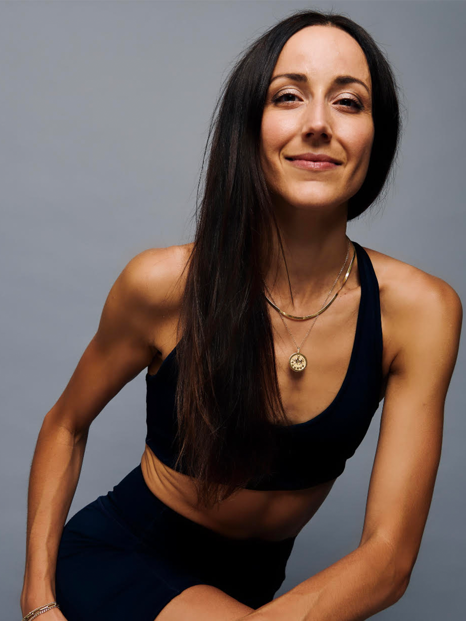 Woman with long dark hair wearing a black sports bra and necklace poses in front of a plain grey background, smiling slightly at the camera.