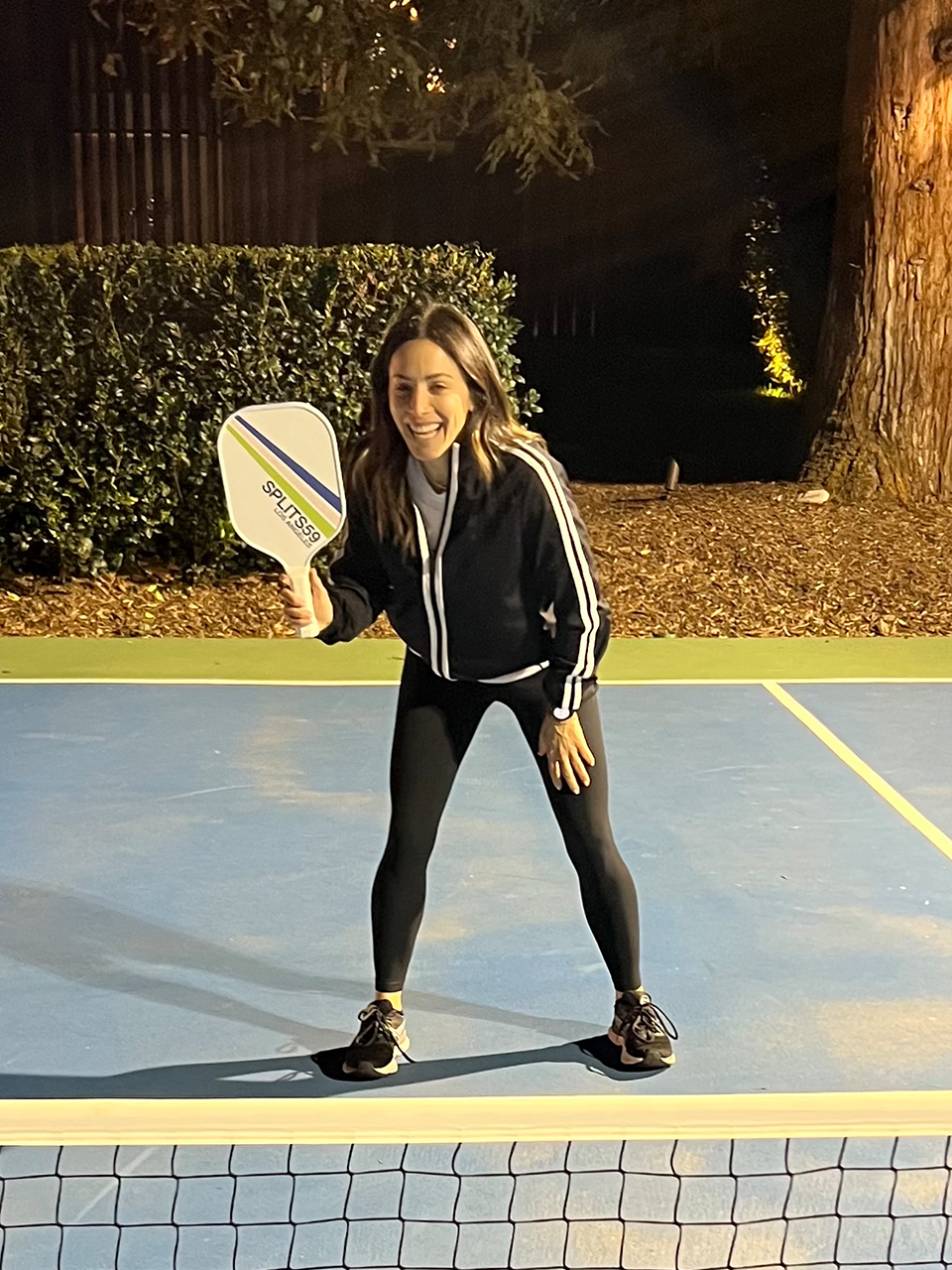 A woman in athletic clothing stands on a pickleball court at night, holding a paddle and smiling, ready to play.