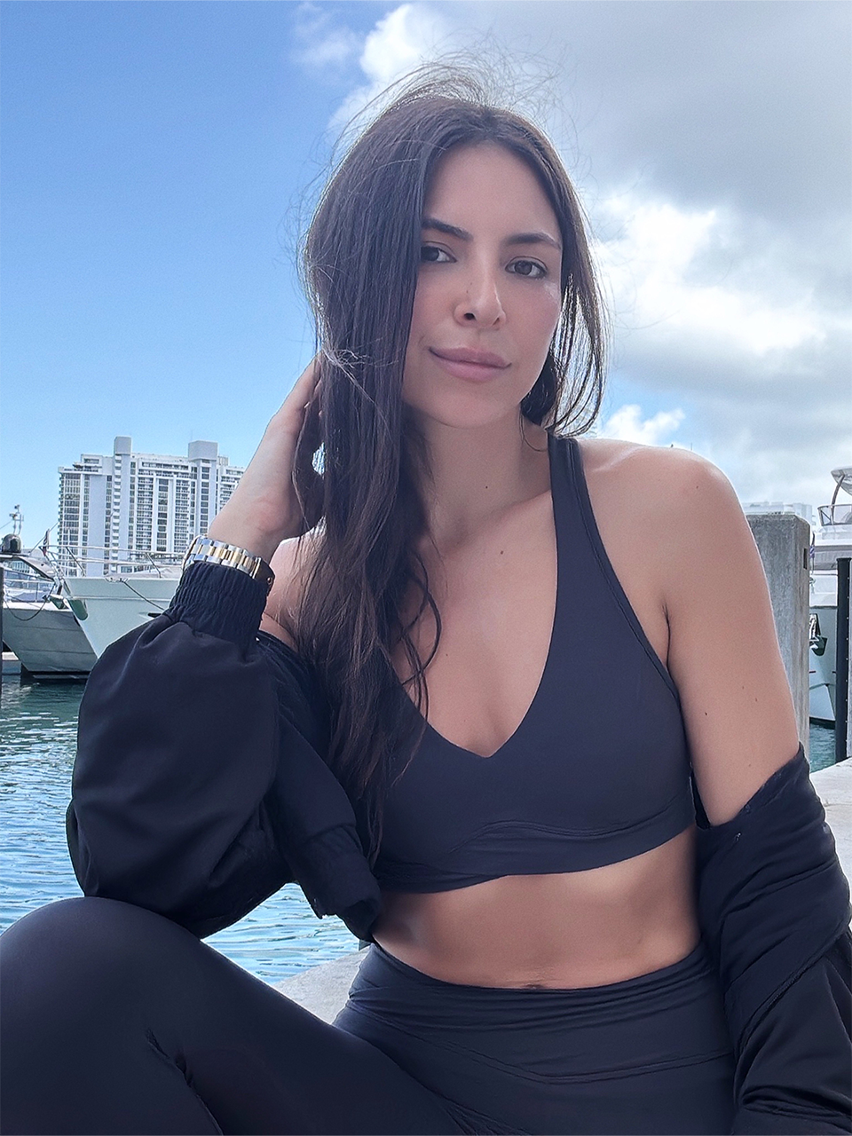 A woman in black athletic wear sits near a marina with boats and tall buildings in the background, under a partly cloudy sky.