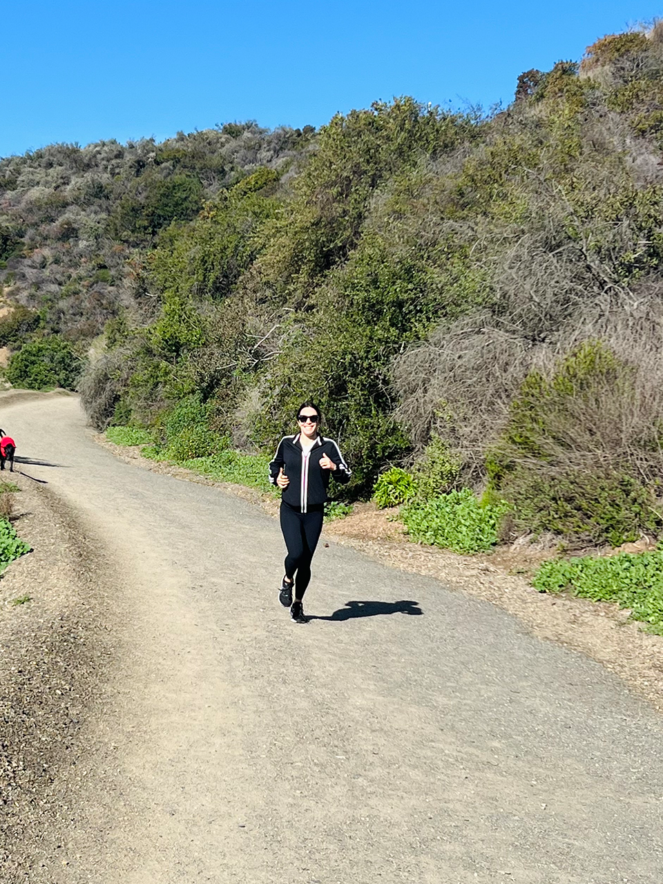 A person in black athletic clothing jogs on a dirt trail surrounded by hills and green shrubs under a clear blue sky.