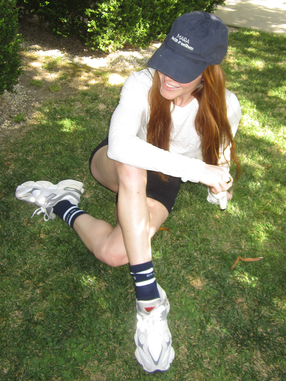 A person with long red hair in a white shirt, black shorts, striped socks, white sneakers, and a navy cap sits on grass with one arm resting on their knee, smiling.
