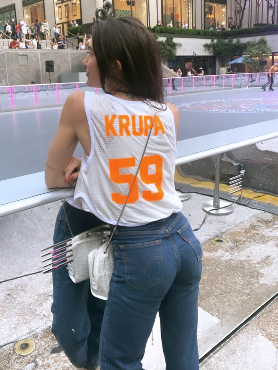A woman wearing a sleeveless jersey with "KRUPA 59" stands by a barrier near an outdoor skating rink, holding a white purse with colored markers.