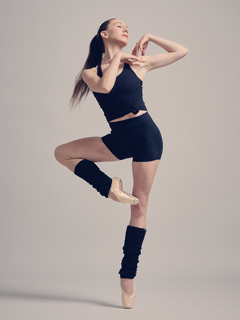 A ballet dancer in black attire and leg warmers poses en pointe with one leg raised and arms gracefully positioned, against a plain background.