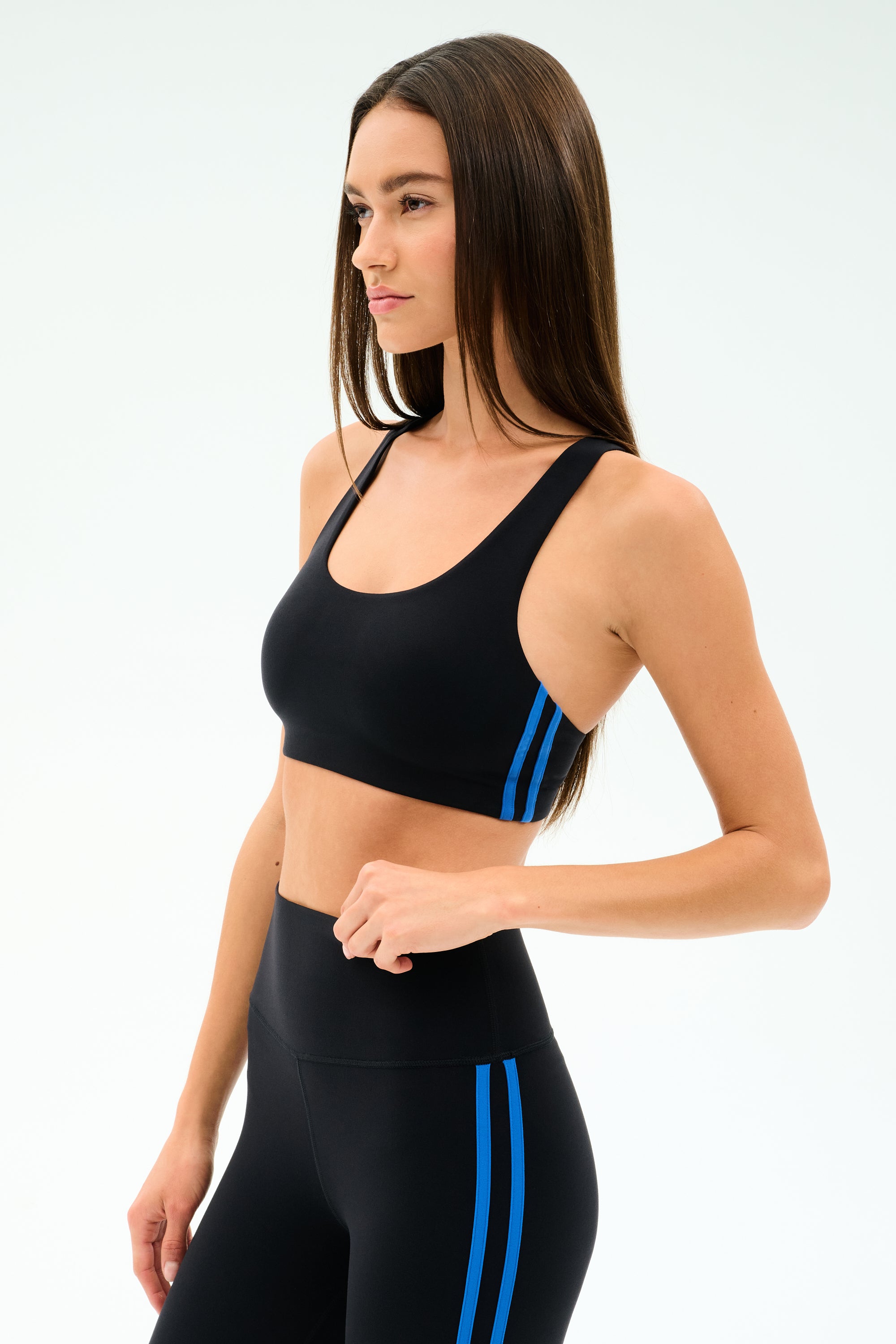 A woman wears the SPLITS59 Ella Airweight Bra in Black/Classic Blue, featuring soft nylon-spandex fabric, paired with high-waisted leggings, standing against a plain white background.
