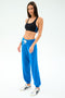 A woman in a black spandex sports bra, SPLITS59 Andie Oversized Fleece Sweatpant in Classic Blue with a white drawstring, and white sneakers stands against a plain light background.