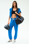 A woman in blue sweatpants and a black sports bra holds the SPLITS59 MZW x Splits59 Large Accordion Cosmetic bag, paired with a small quilted black bag and white sneakers.