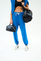 A person in blue sweatpants and a black sports bra holds two SPLITS59 MZW x Splits59 Large Accordion Cosmetic bags, with a blue jacket over their shoulders and white sneakers.