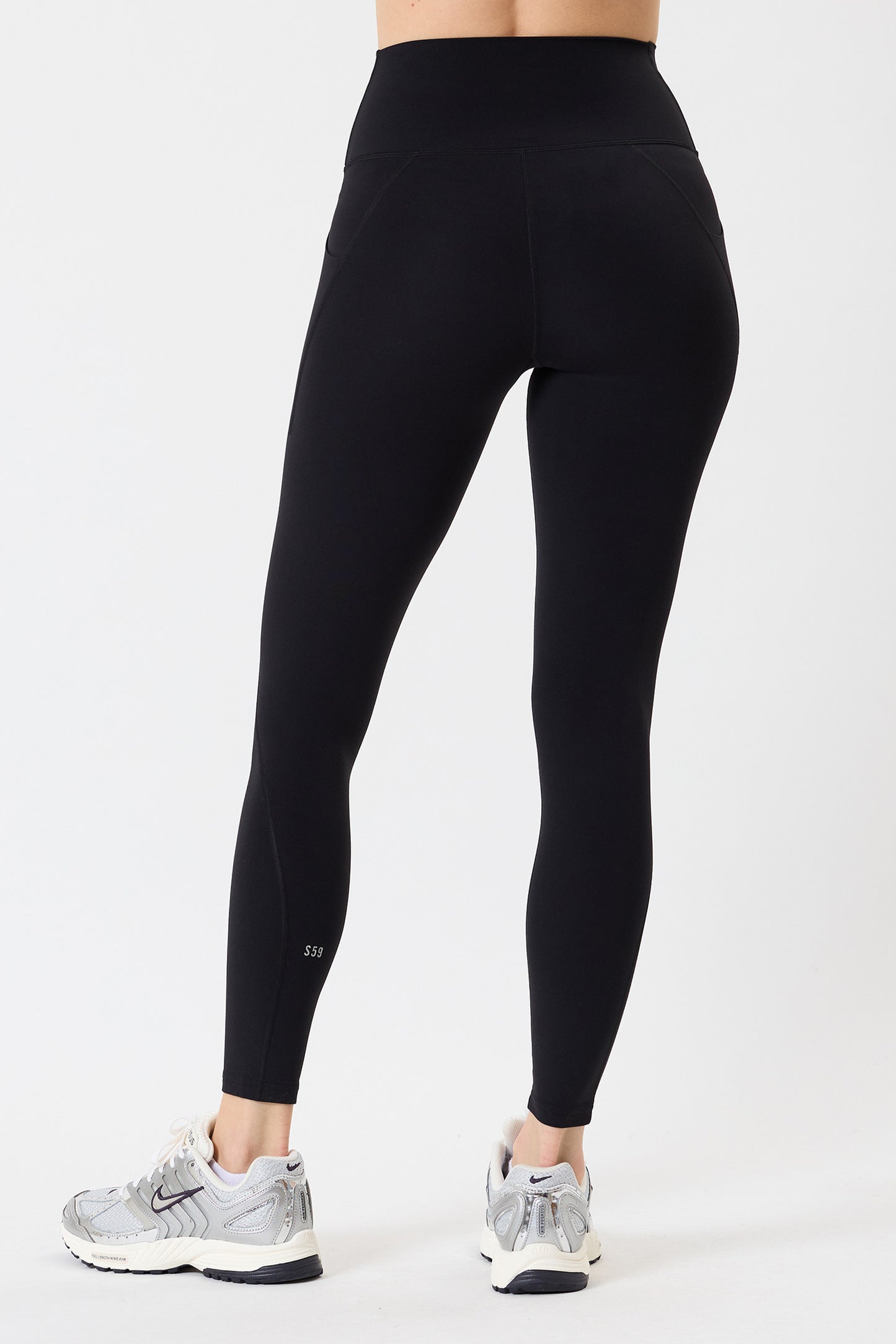 A person wearing SPLITS59 Airweight High Waist 7/8 Pocket Legging in black and gray athletic shoes stands facing away against a white background.