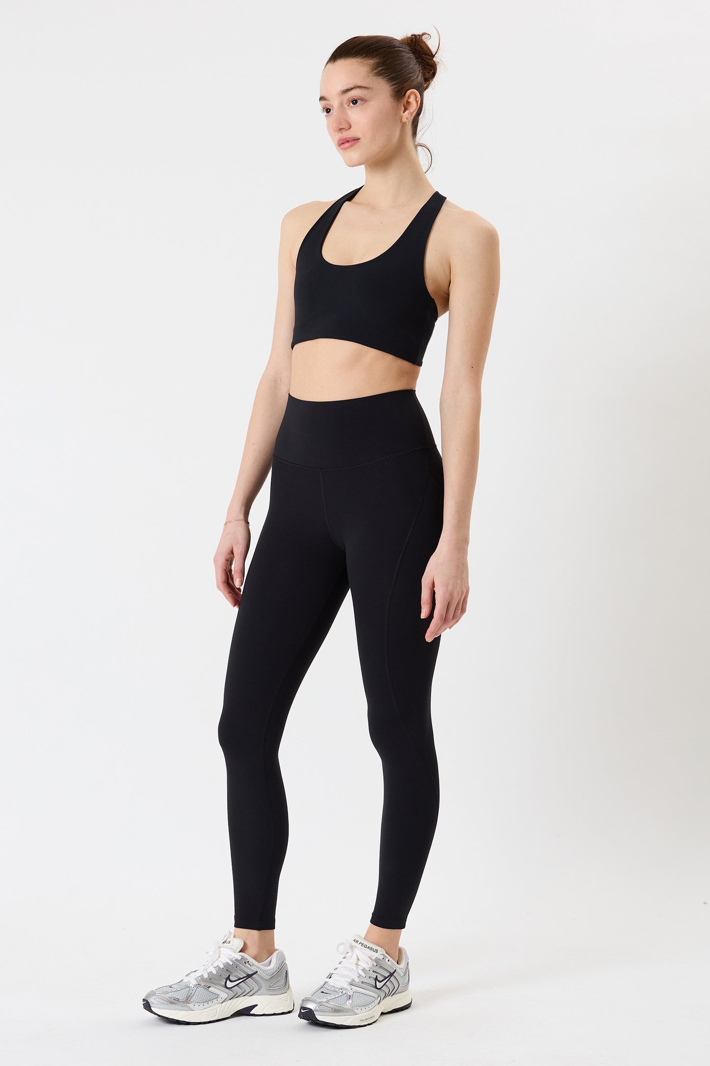 A woman faces forward, wearing a black SPLITS59 Airweight High Waist 7/8 Pocket Legging—ideal for hot yoga—with a black sports bra and gray athletic shoes, standing against a plain white background.