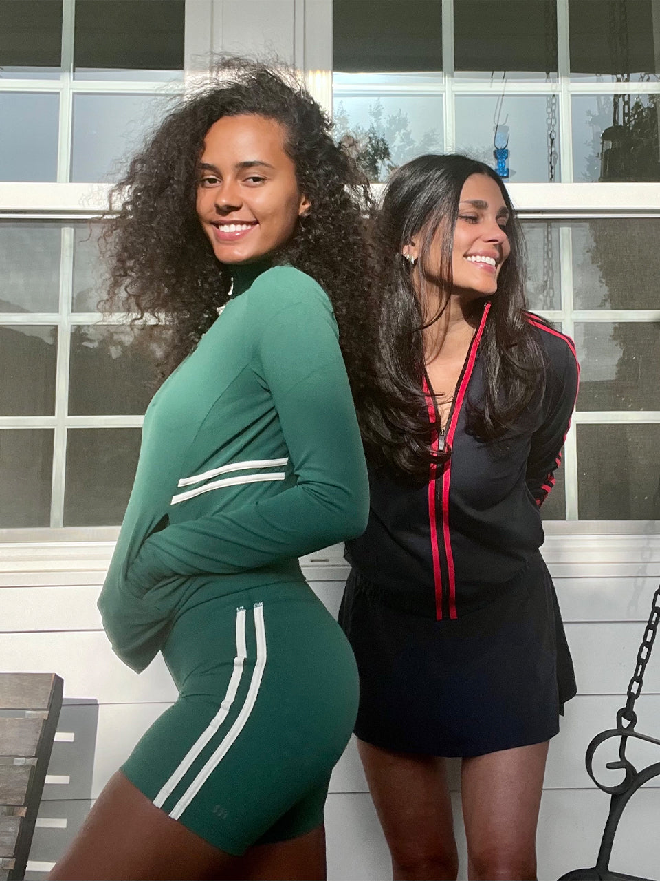 Two women stand together indoors, smiling. One wears a green athletic outfit with white stripes, the other wears a black dress with red details. They are in front of large window panes.