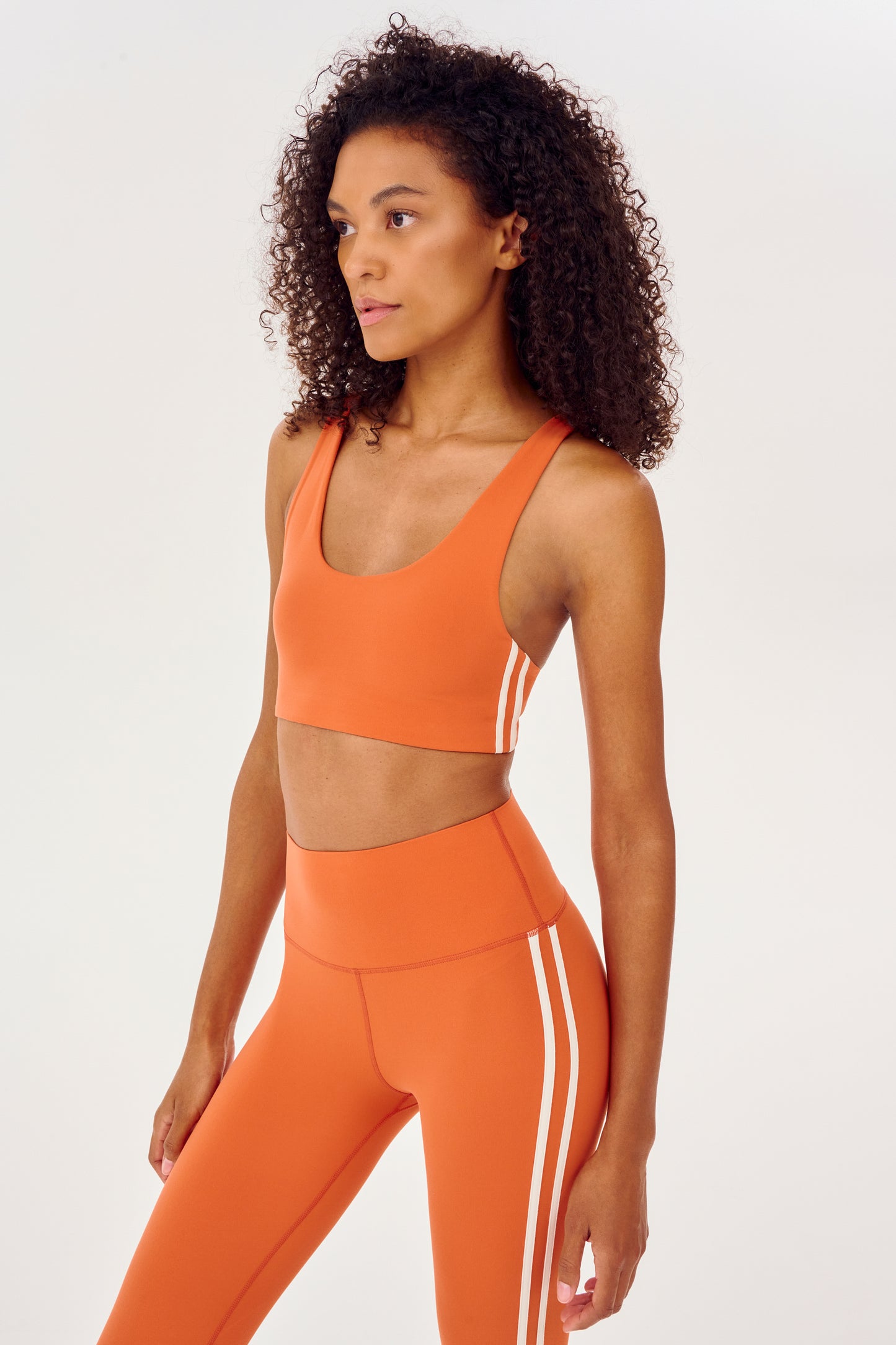 Side view of girl wearing orange sports bra with two thin white stripes down the side and orange leggings