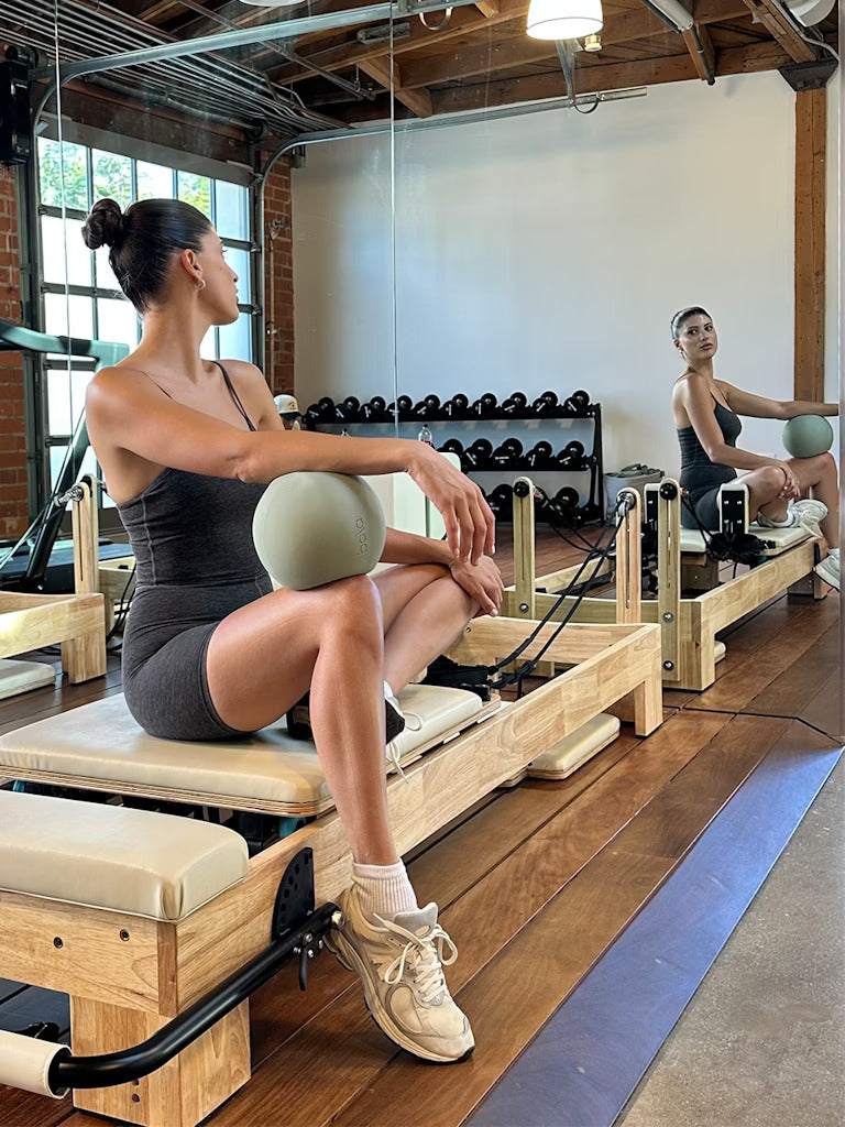 A woman in athletic wear sits on a Pilates reformer, holding a small exercise ball, and looks toward her reflection in a gym with wooden floors and exposed brick walls.